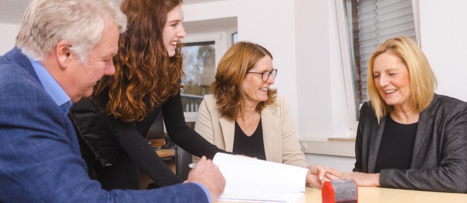Vier Personen schauen gemeinsam in ein Dokument und lachen dabei. Die Atmosphäre wirkt freundlich und professionell