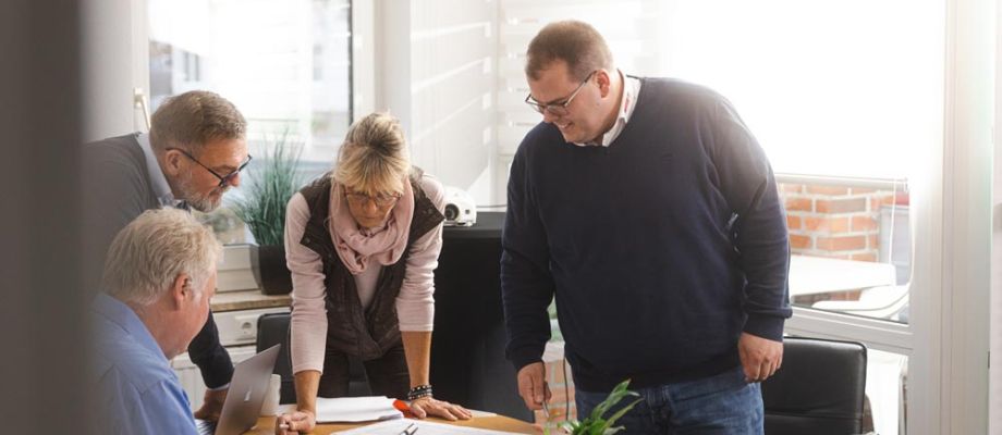 Eine Gruppe von vier Personen, arbeitet gemeinsam in einem modernen Büro. Sie stehen und sitzen um einen Tisch mit Bauplänen, Laptops und Notizen.