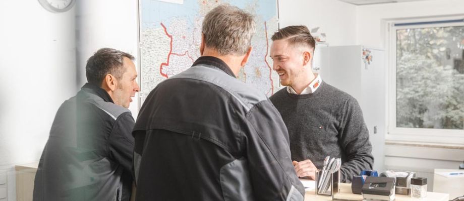 Drei Männer stehen in einem Büro und unterhalten sich freundlich. Zwei von ihnen tragen dunkle Arbeitskleidung, während der dritte in einem grauen Pullover gekleidet ist.