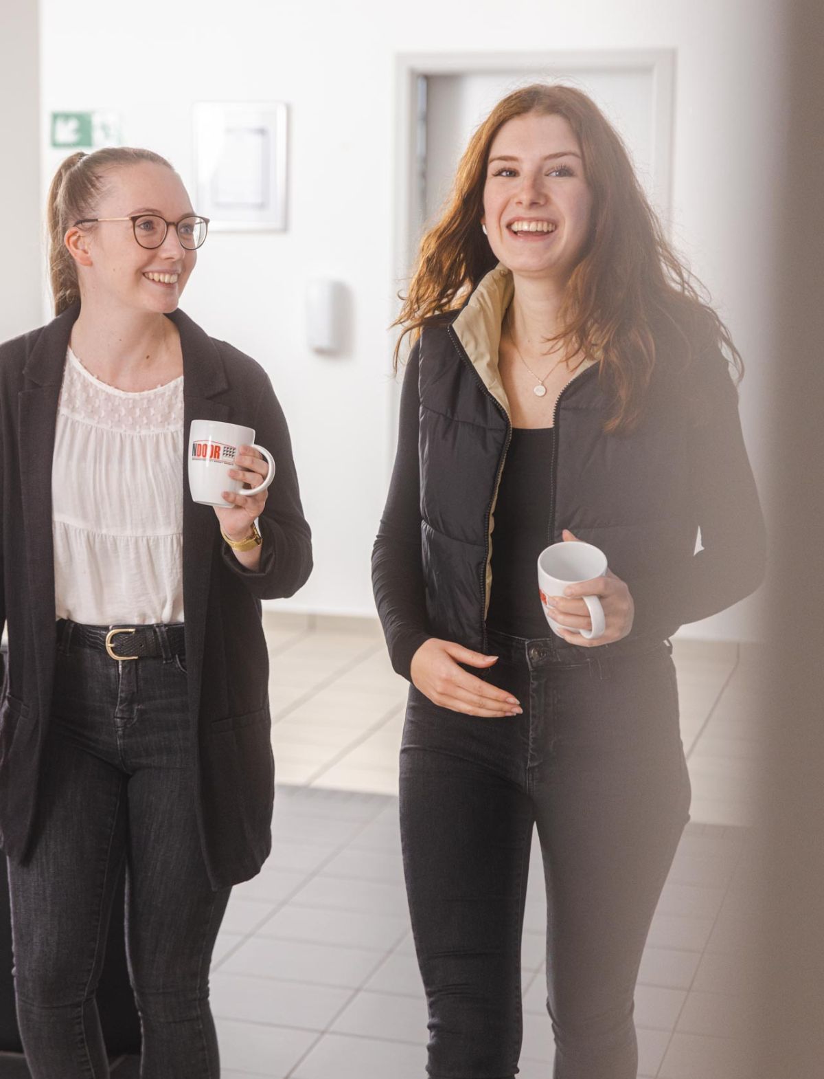 Zwei junge Frauen in einem modernen Bürogebäude unterhalten sich lachend, während sie jeweils eine Kaffeetasse mit 'NDOOR'-Logo in der Hand halten.