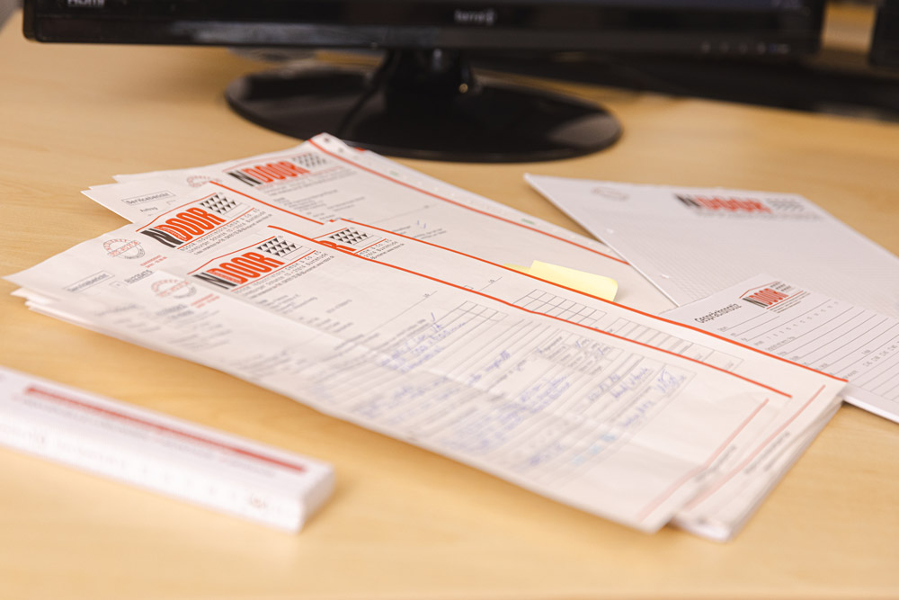 Mehrere ausgefüllte Auftragsformulare und Dokumente der Firma NDOOR liegen auf einem Schreibtisch, im Hintergrund ist ein Monitor zu sehen.