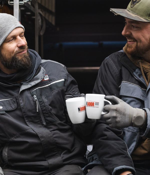Zwei Männer in dunkler Arbeitskleidung und Handschuhen sitzen entspannt nebeneinander und stoßen mit weißen Kaffeetassen an, auf denen 'NDOOR' steht.