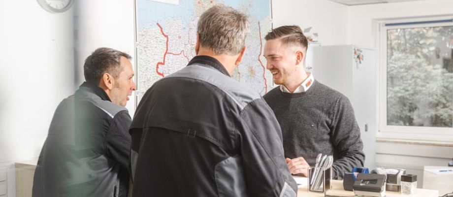 Drei Männer stehen in einem Büro und unterhalten sich freundlich. Zwei von ihnen tragen dunkle Arbeitskleidung, während der dritte in einem grauen Pullover gekleidet ist.