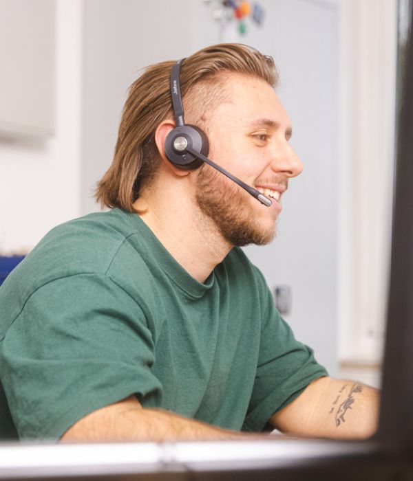 Ein junger Mann mit halblangen Haaren, Bart und einem Headset sitzt vor einem Computerbildschirm und lächelt