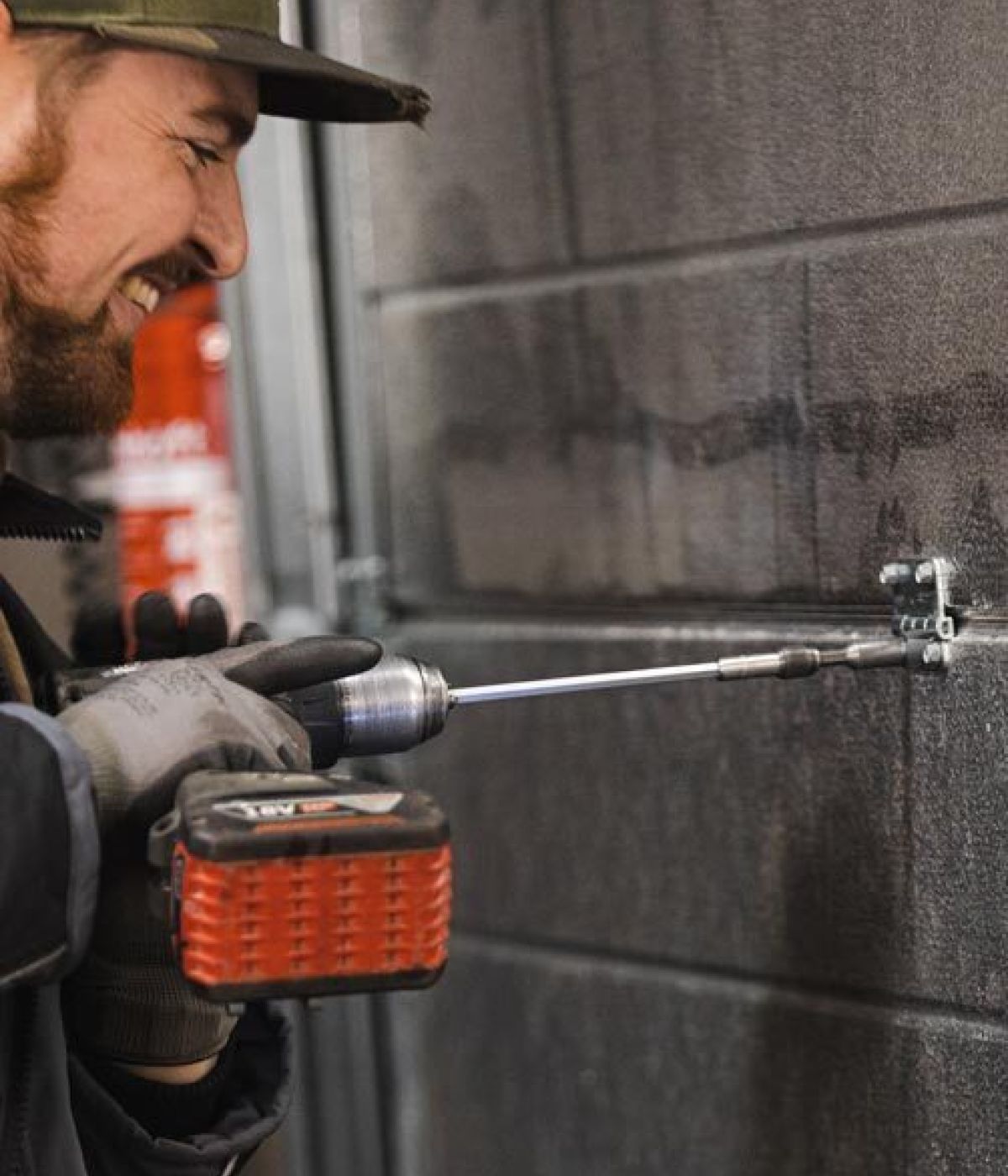 Handwerker mit Arbeitshandschuhen befestigt mit einer Akkubohrmaschine eine Halterung an einer dunklen Metalltür und lächelt dabei.