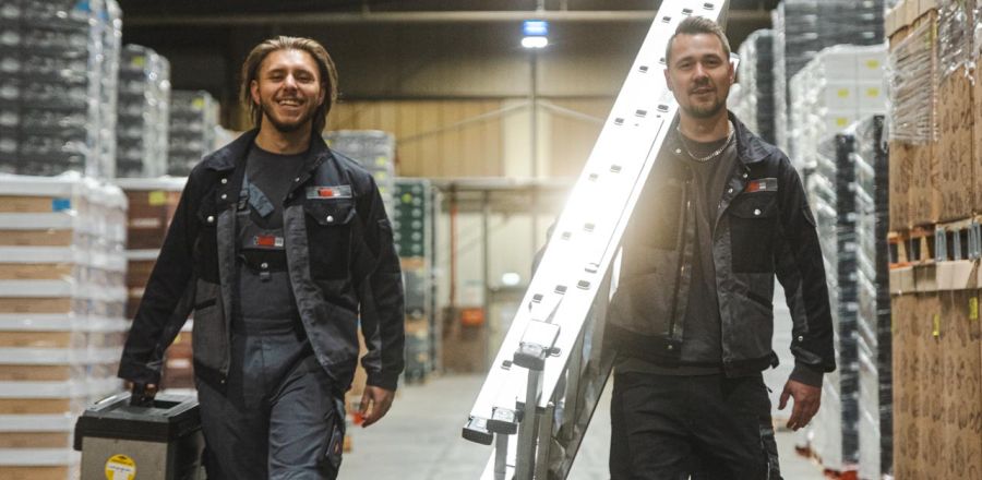 2 Mitarbeiter in einer Halle mit Leiter und Werkzeugkoffer in der Hand.