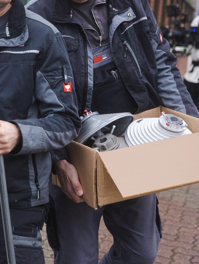 Zwei Handwerker in dunkler Arbeitskleidung tragen einen Karton mit Bauteilen, vermutlich für eine Garagentorinstallation.