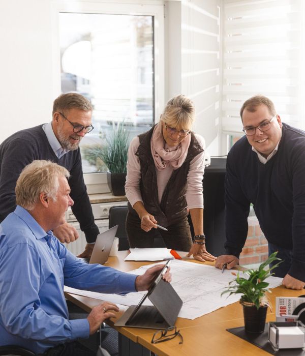Eine Gruppe von vier Personen, arbeitet gemeinsam in einem modernen Büro. Sie stehen und sitzen um einen Tisch mit Bauplänen, Laptops und Notizen. 