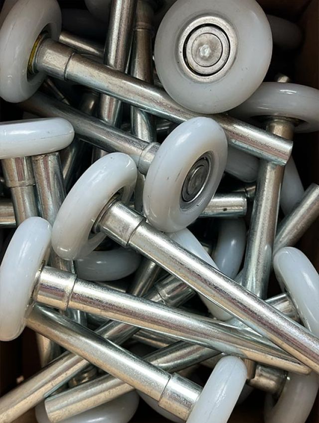 Mehrere silberne Laufrollen mit weißen Kunststoffrädern, vermutlich für Garagentore oder Schienensysteme, in einer Kiste.